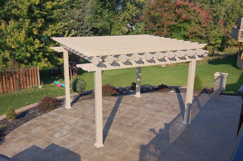 White pergola on a stone patio in a backyard, surrounded by trees and greenery.