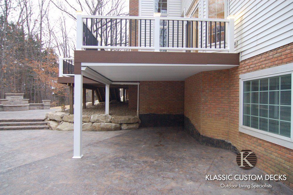 Covered patio under a white deck beside a brick house, with stone steps and retaining wall.