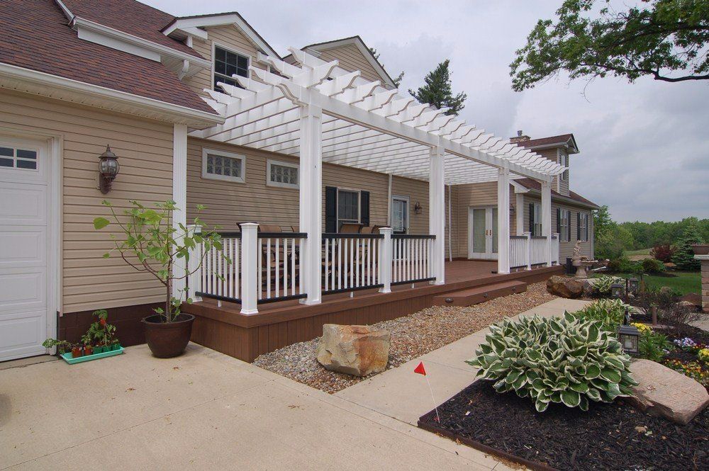 Backyard patio with white pergola, beige house siding, wooden deck, and landscaped garden beds