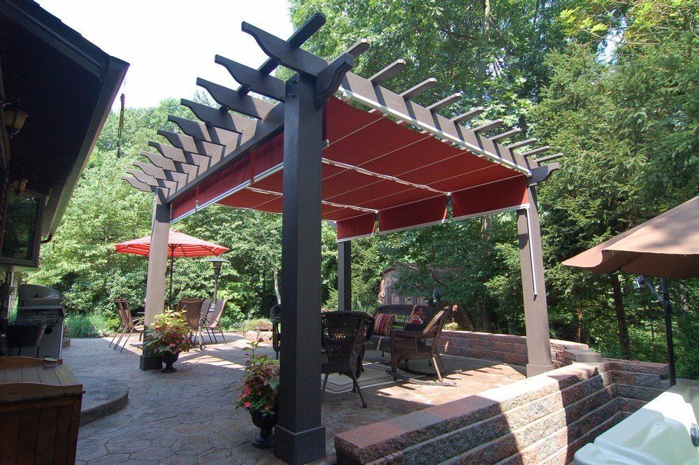 Outdoor patio with red pergola, black support posts, potted plants, and chairs in a wooded garden.