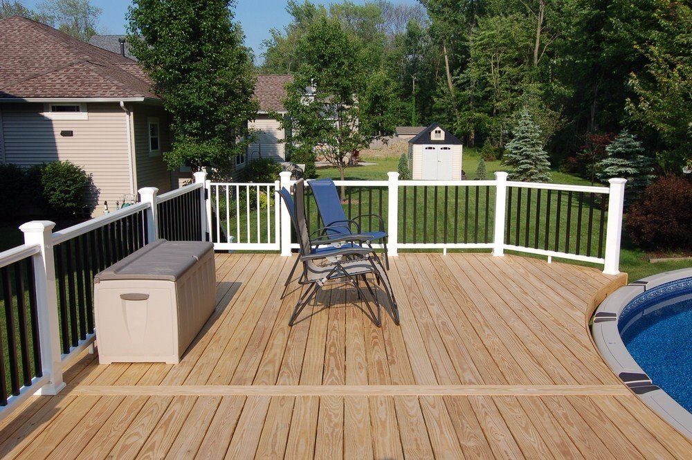 Wooden deck with patio chairs, a storage bench, white railing, and a pool edge in a sunny backyard