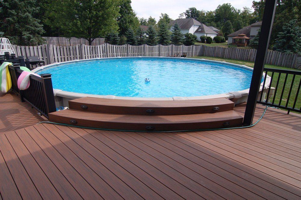 Backyard above-ground pool with wooden deck and steps, surrounded by a fence and trees