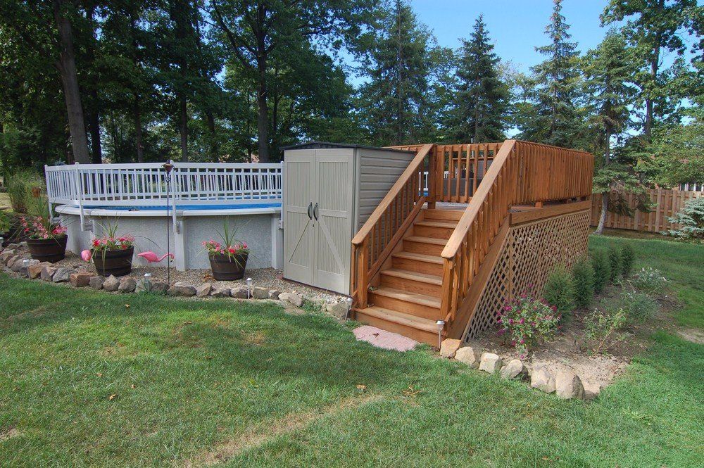 Wooden backyard deck with stairs beside a raised pool and potted plants, surrounded by grass and trees