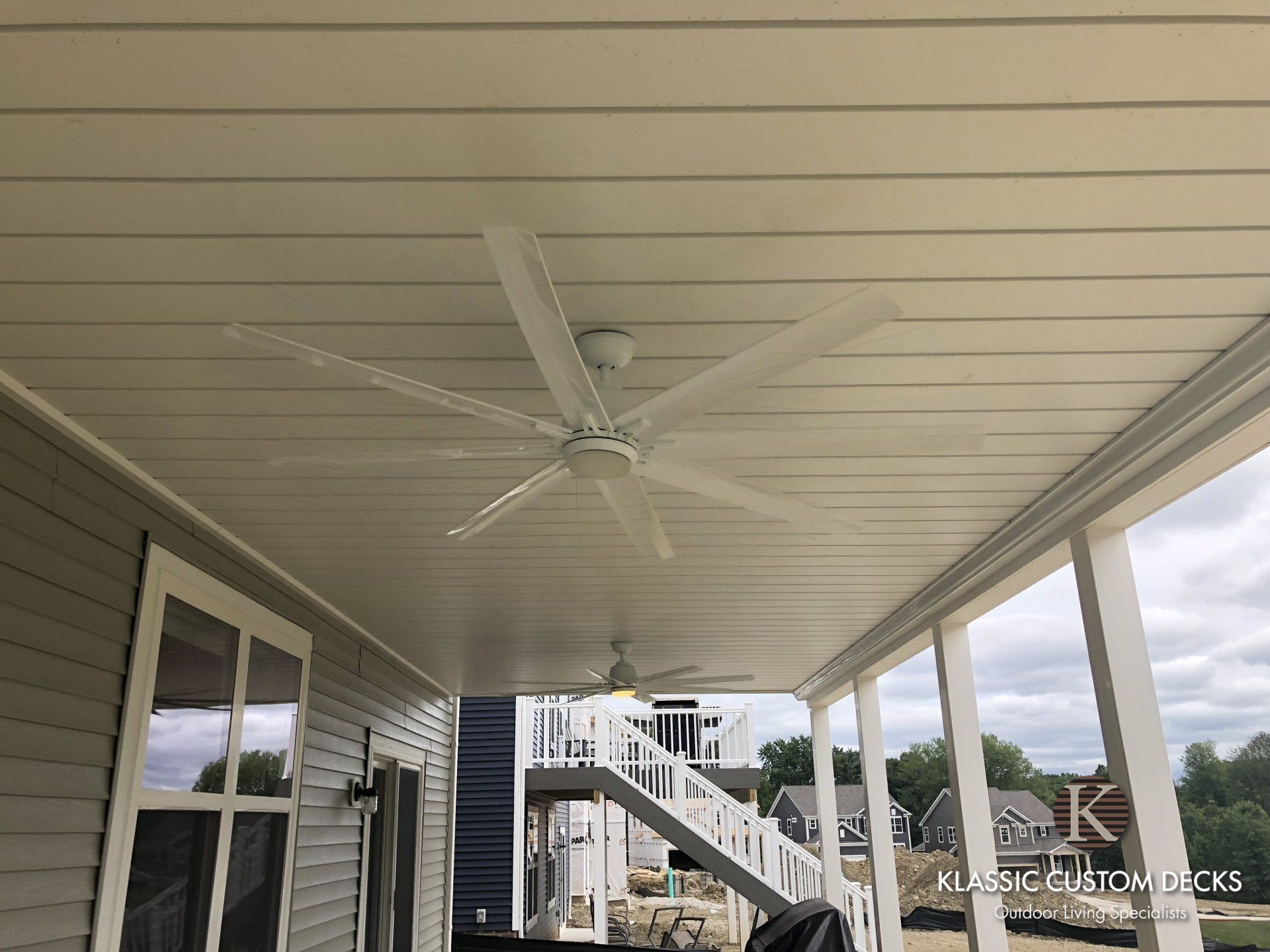 Covered apartment balcony with white ceiling fan, railing, and staircase view beyond.