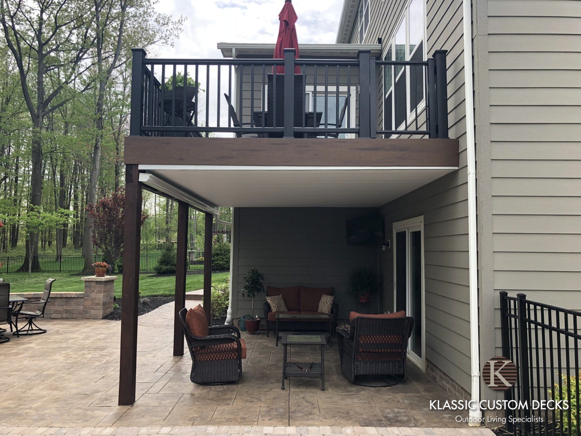 Covered backyard patio under an elevated deck with outdoor seating and a red umbrella