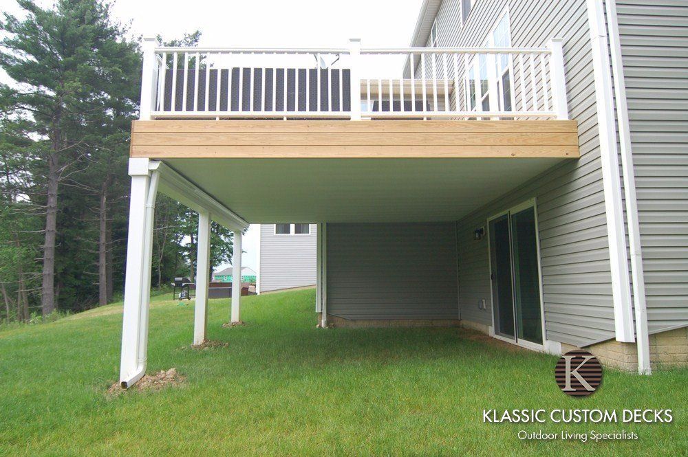Raised wooden deck over a shaded patio beside a house, with white railing and support posts.