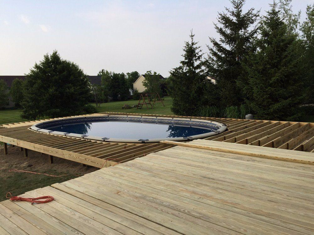 Wooden deck under construction around an oval above-ground pool in a grassy yard with trees.