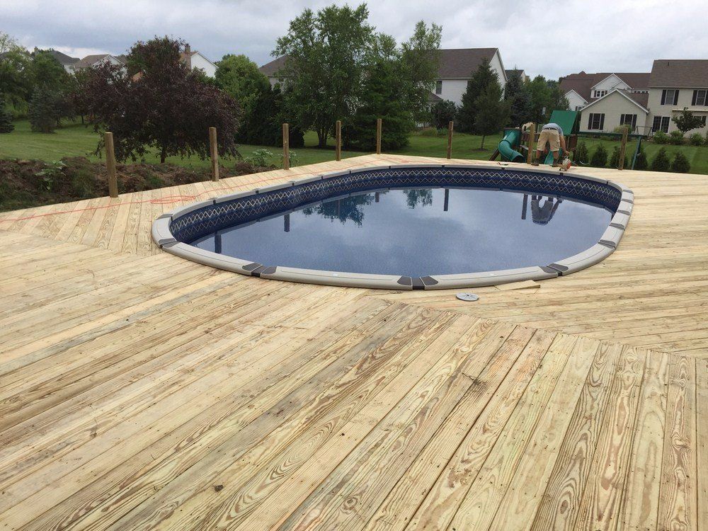 Oval backyard swimming pool with dark water on a wooden deck, surrounded by trees and houses