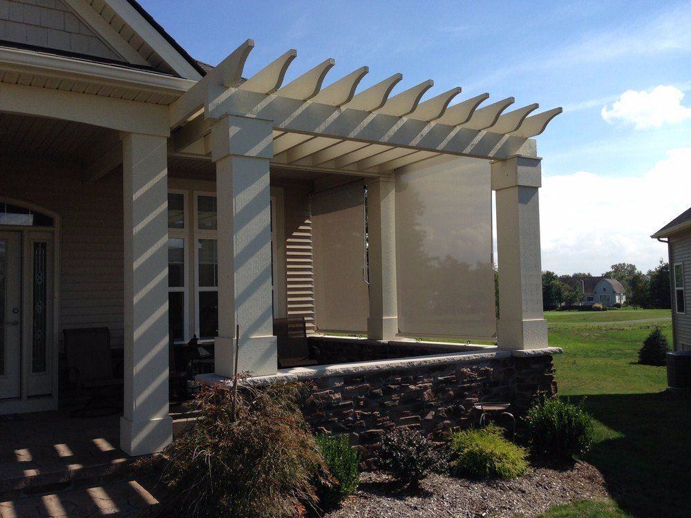House porch with white pergola beams, stone base, and window shades overlooking a green lawn.