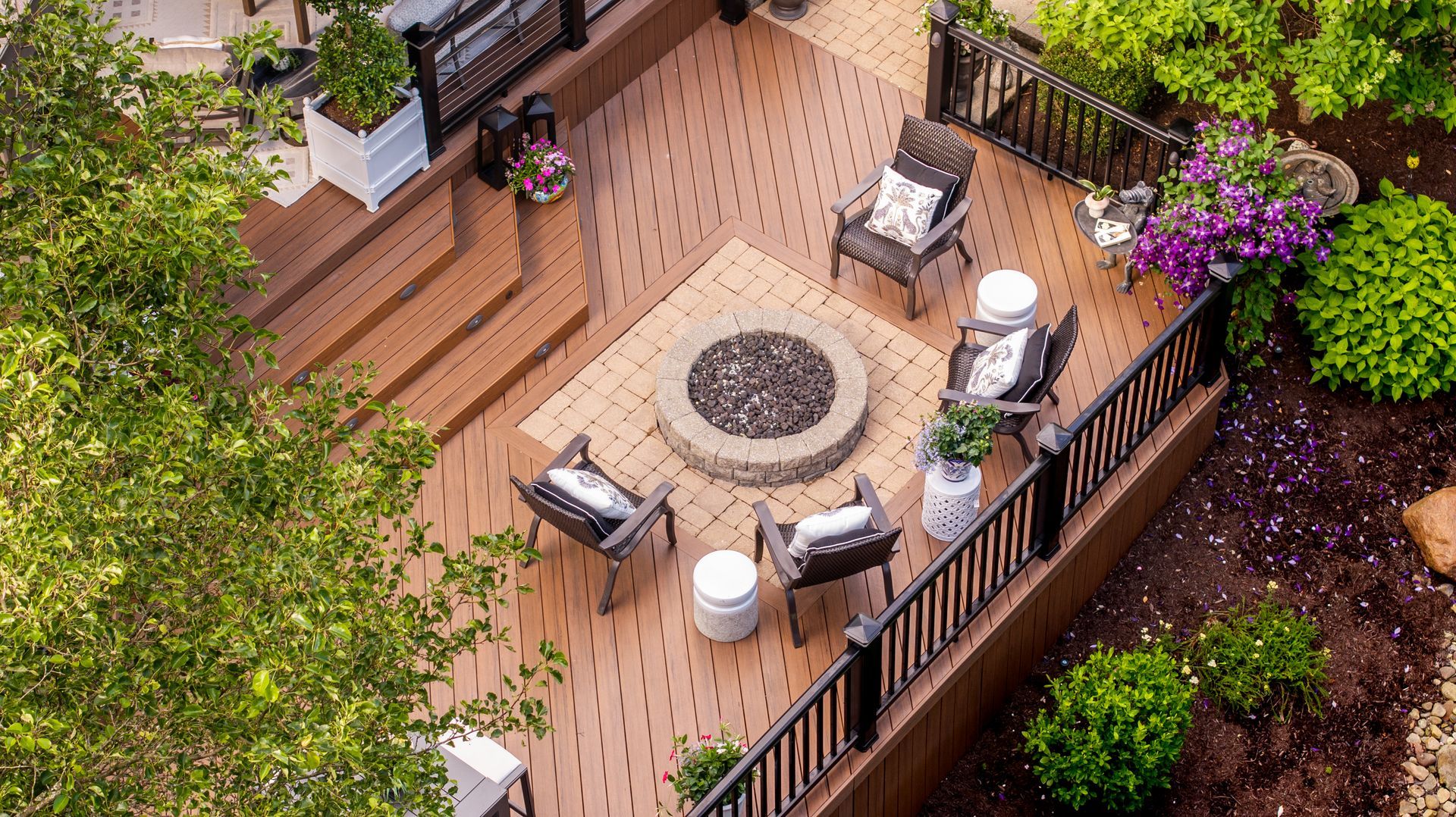 Wooden deck with patio furniture around a fire pit, bordered by a railing and surrounded by greenery.