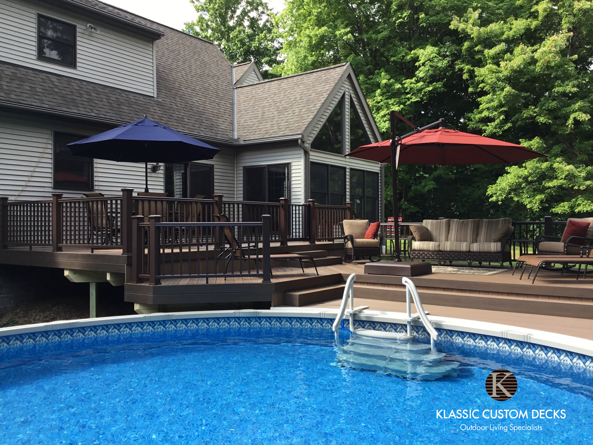 Backyard deck and blue swimming pool with umbrellas, patio furniture, and a house in the background