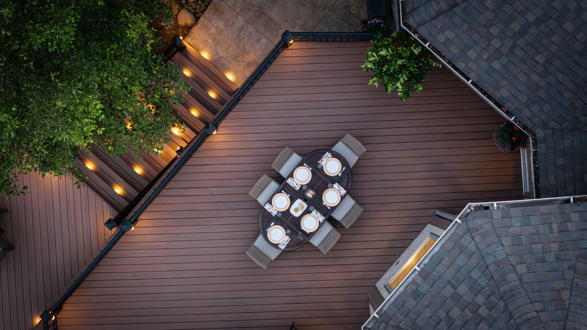 Overhead view of a wooden rooftop deck with an outdoor dining table, chairs, and warm string lights along the edge