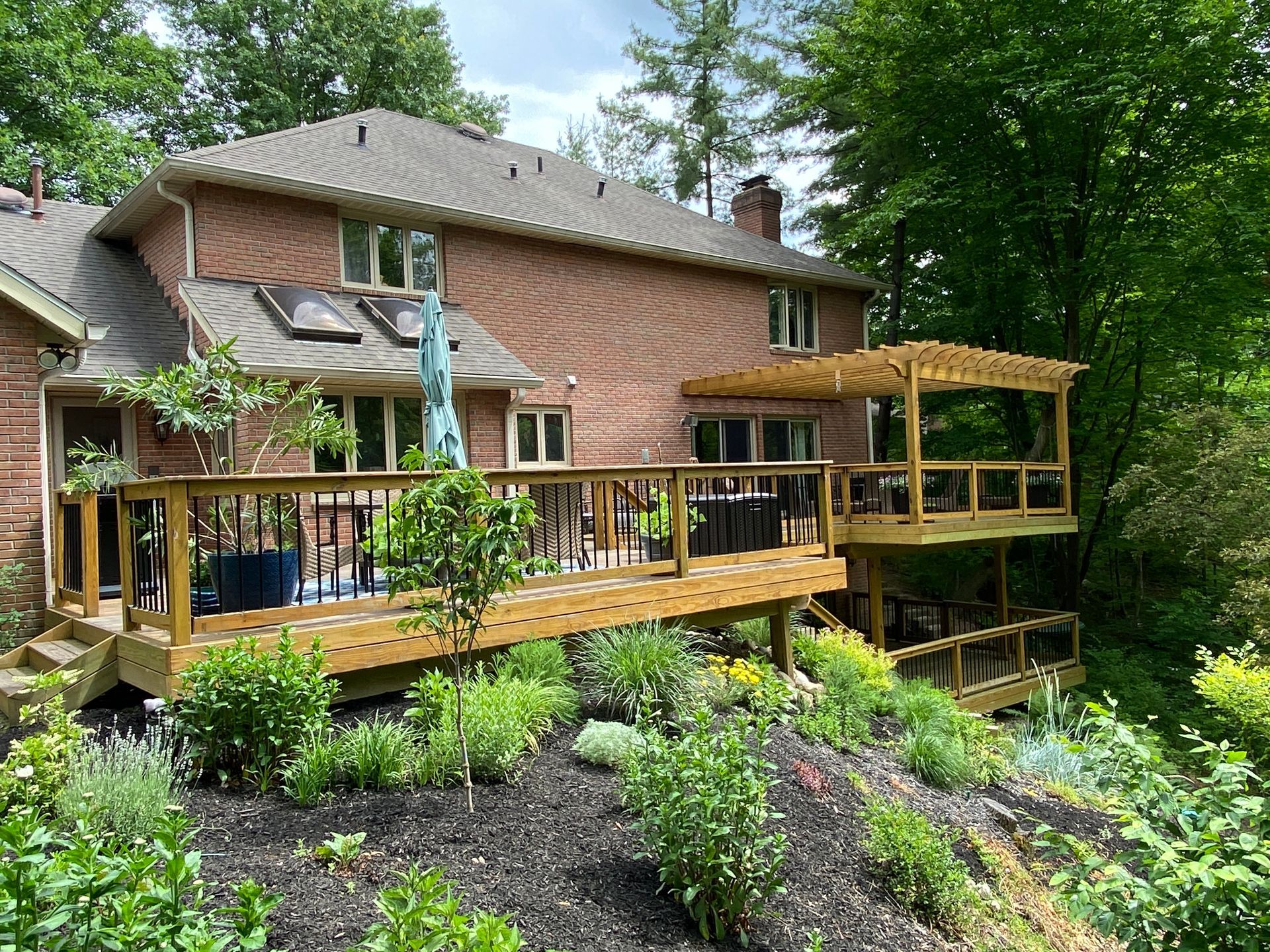 Brick house with a wooden deck overlooking a lush green garden and wooded backyard.