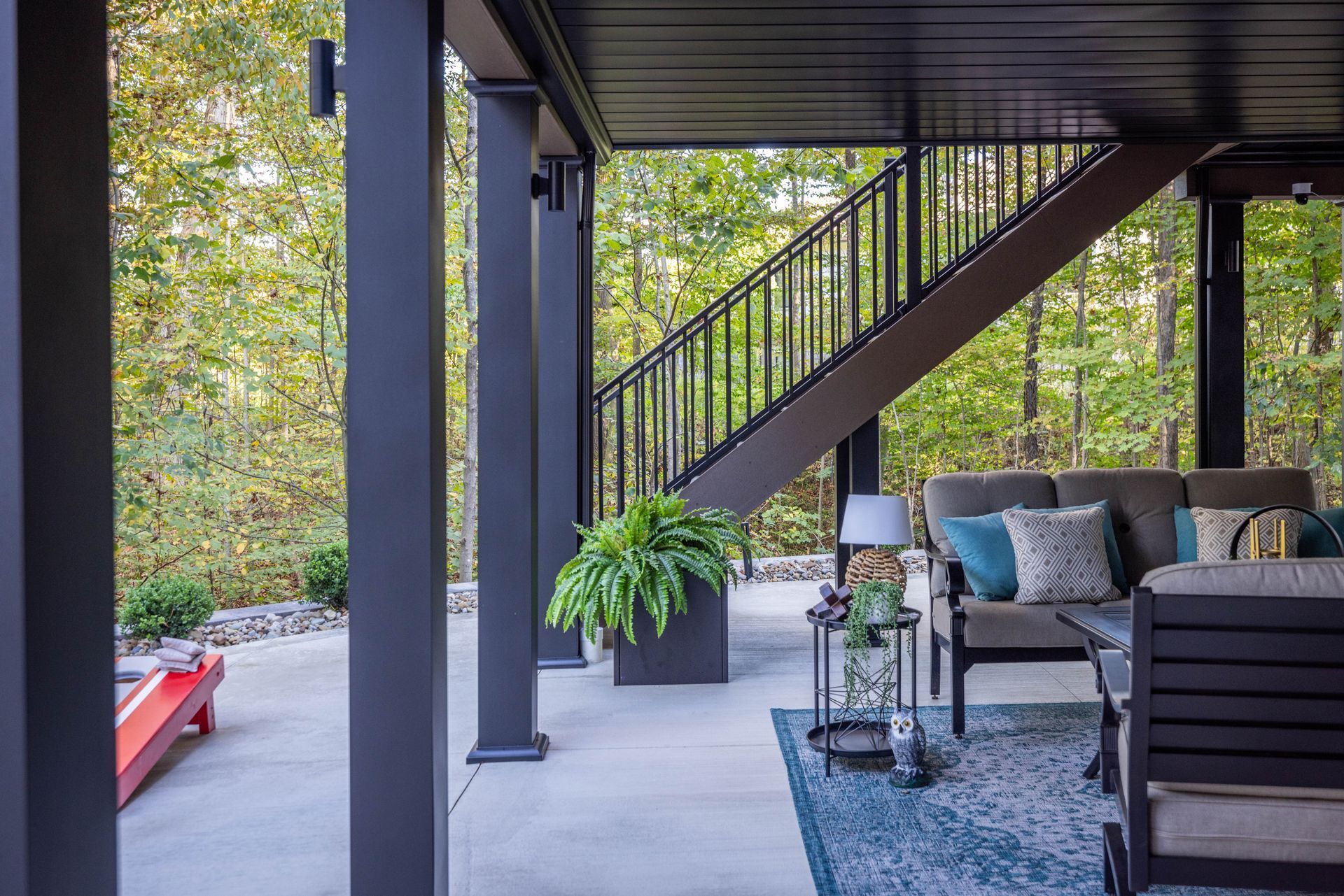 Covered patio with modern outdoor seating, staircase, and a potted plant beside a wooded view.