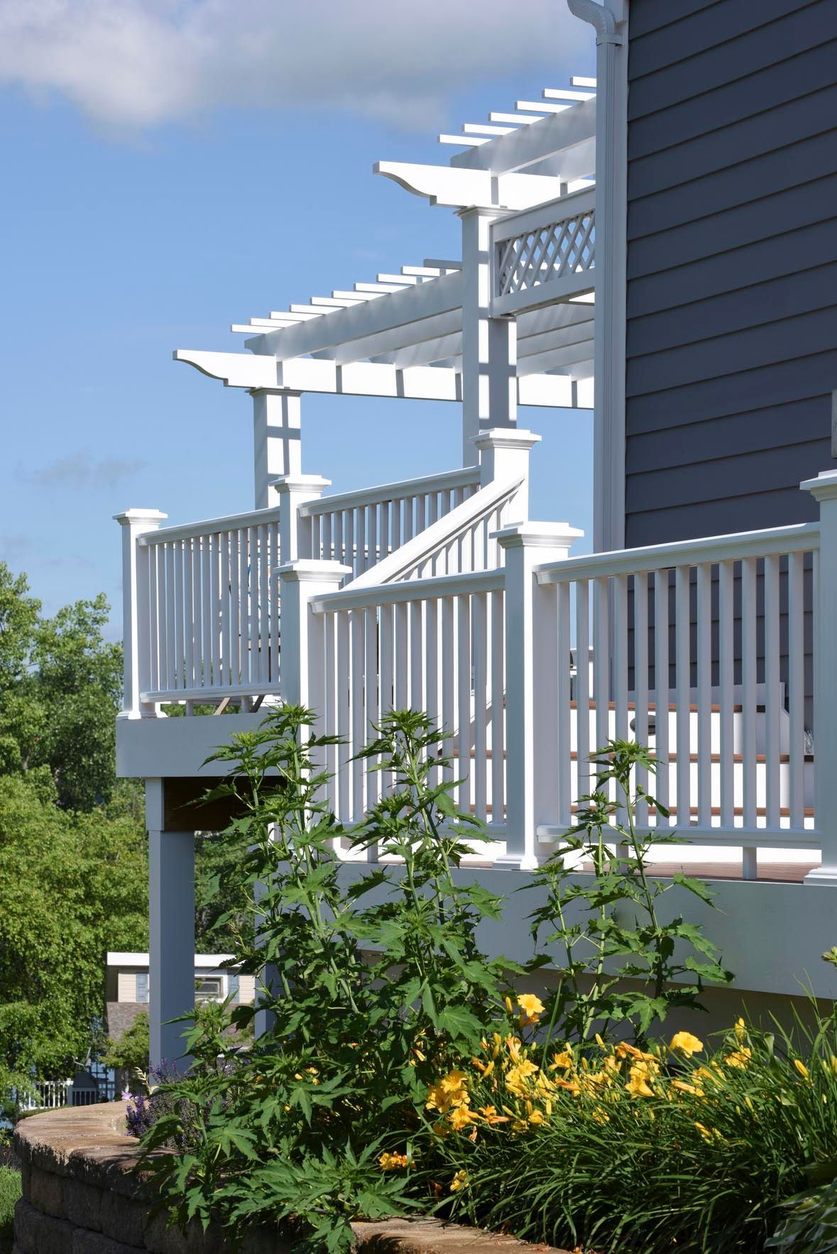 White porch with pergola railings beside a dark blue house, overlooking a garden of green shrubs and yellow flowers
