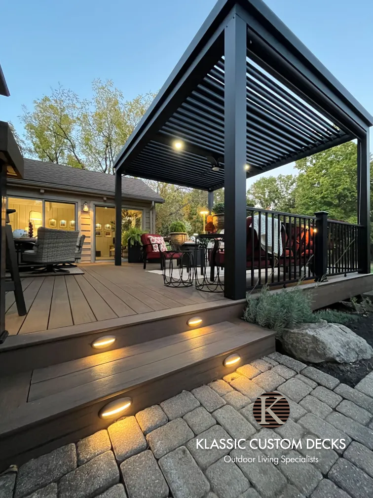 Modern backyard deck with pergola, string lights, patio seating, and steps beside a stone paver patio.