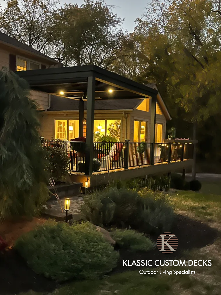 Outdoor patio at dusk with warm lights, glass railings, and landscaped garden beside a house.