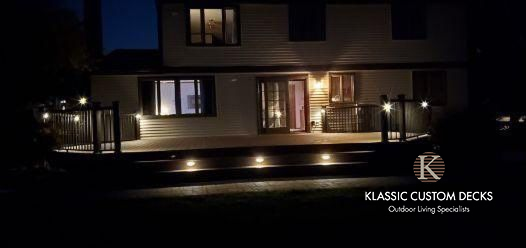 Nighttime view of a lit two-story house with a deck and stone patio, branded “Klassic Custom Decks” in the corner