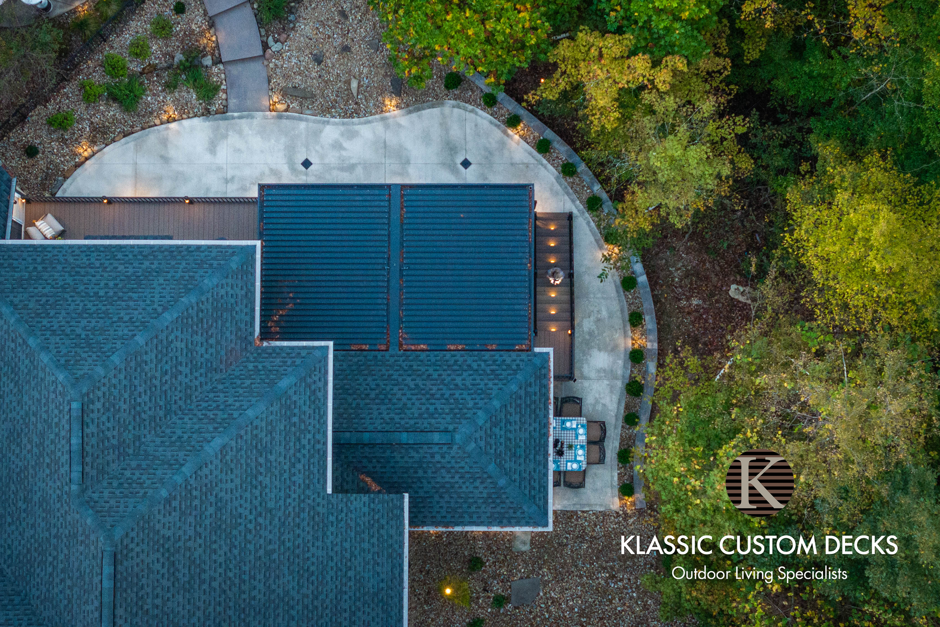 Aerial view of a blue-roofed house with a large deck beside trees and gravel driveway.