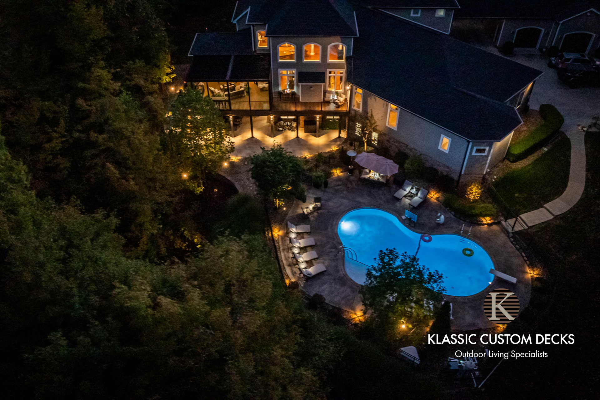 Aerial view of a lit backyard with a house, patio seating, and a glowing blue swimming pool at night