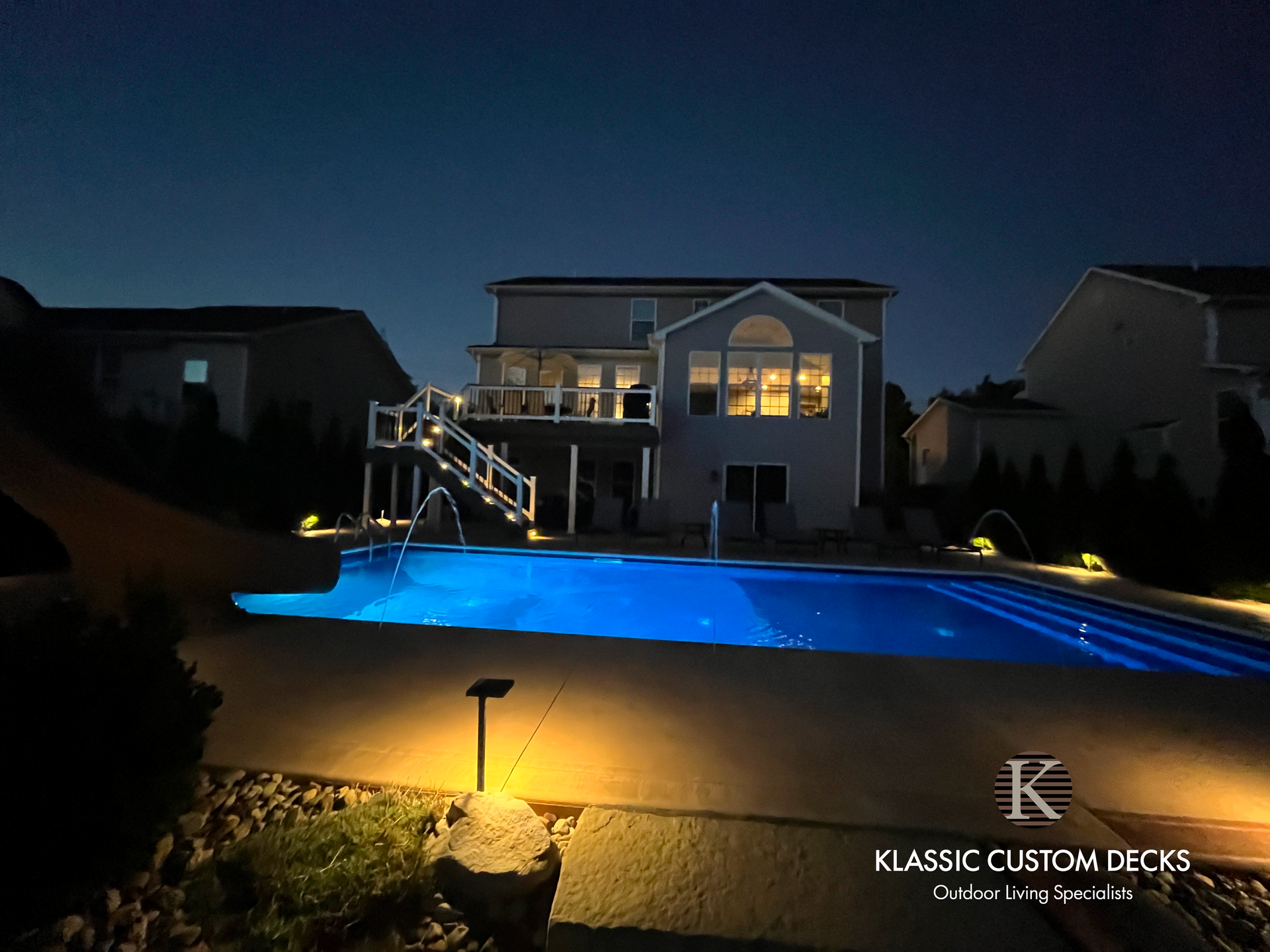 Nighttime backyard with a lit blue pool and a two-story house with glowing windows and deck stairs.
