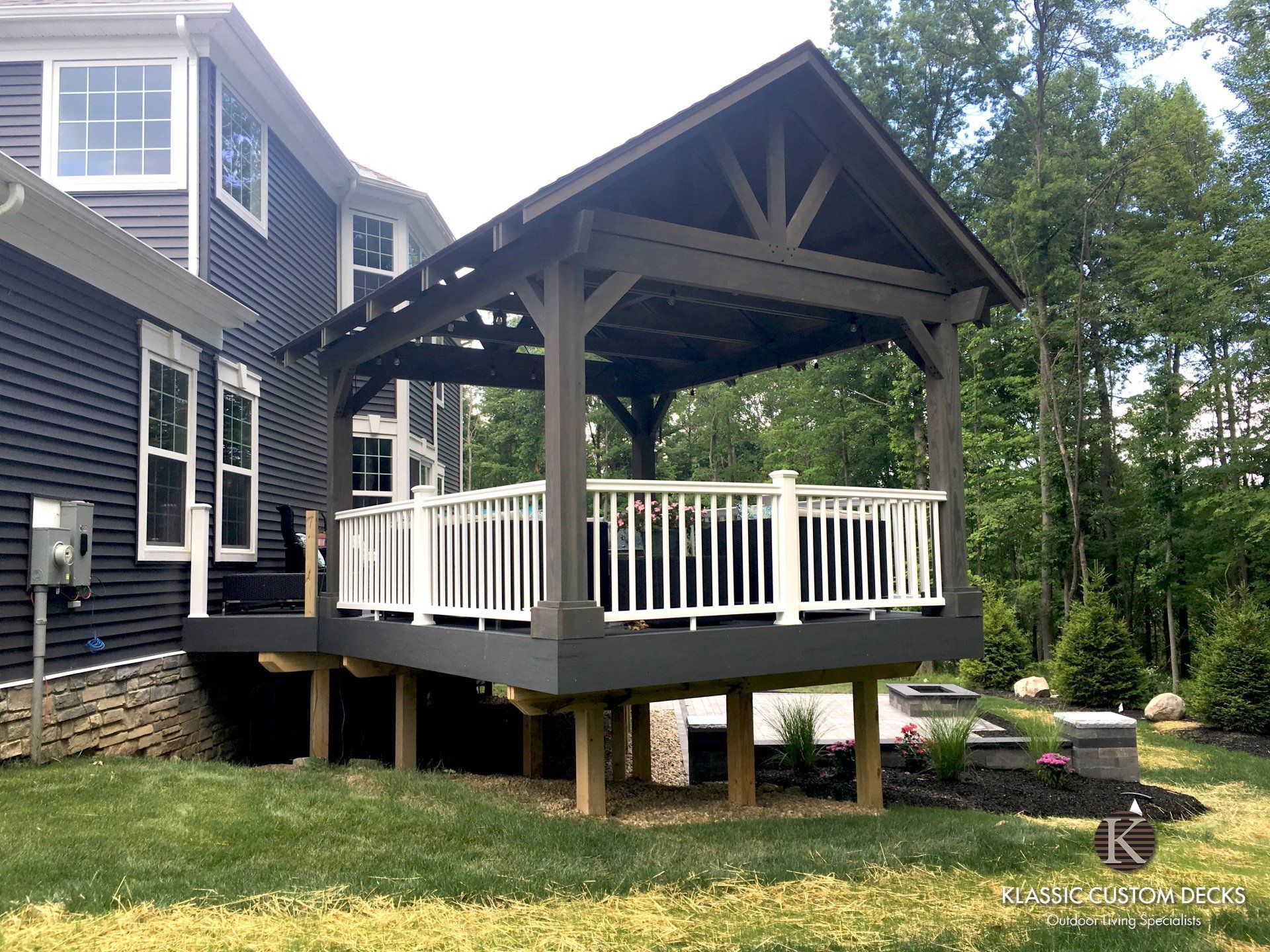 Black raised gazebo deck with white railings beside a gray house, surrounded by trees and lawn.