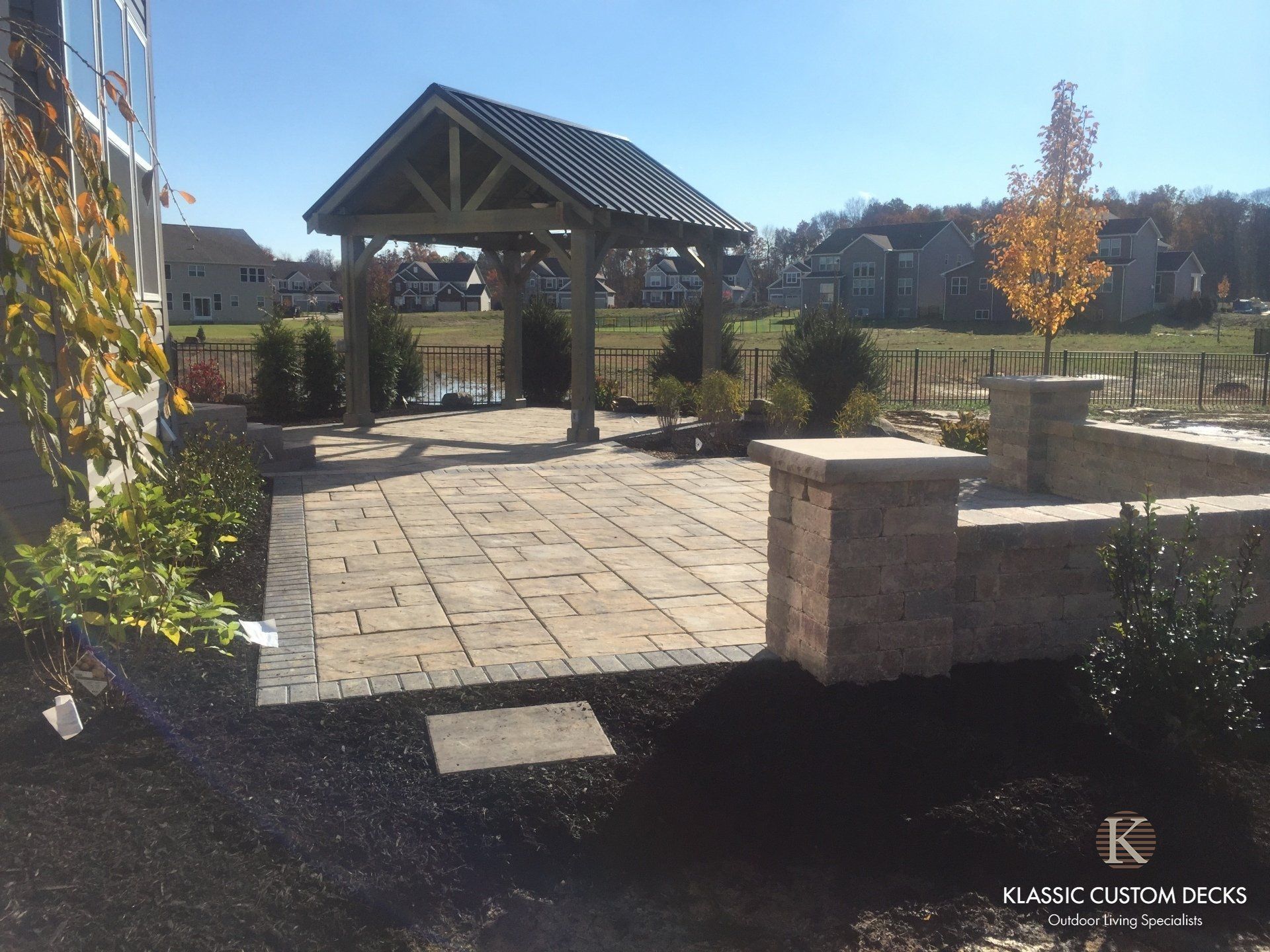Stone patio and covered pavilion in a sunny landscaped park, with benches and autumn trees.