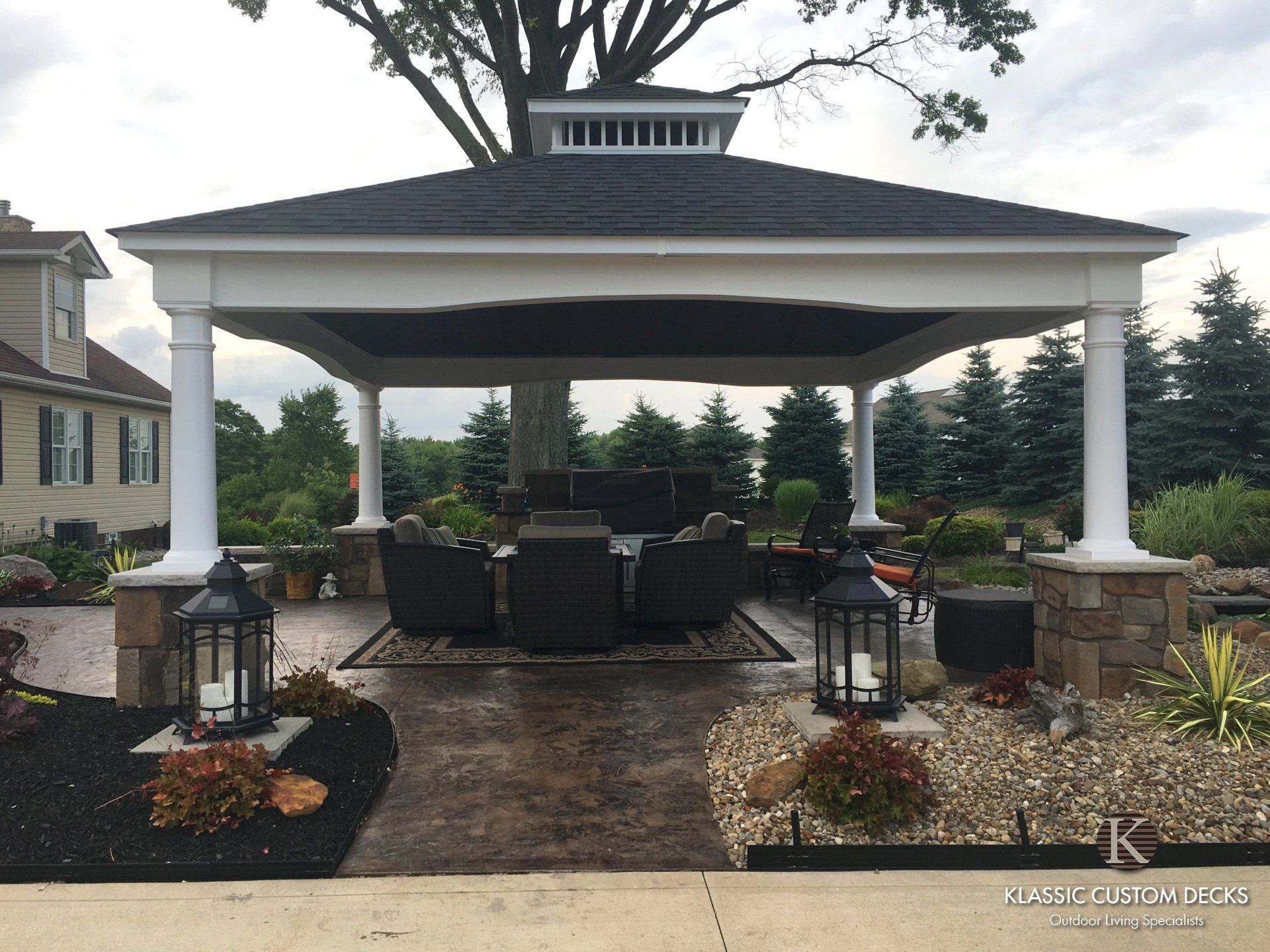 Covered outdoor patio with white columns, dark roof, and wicker seating on a stone walkway in a garden setting