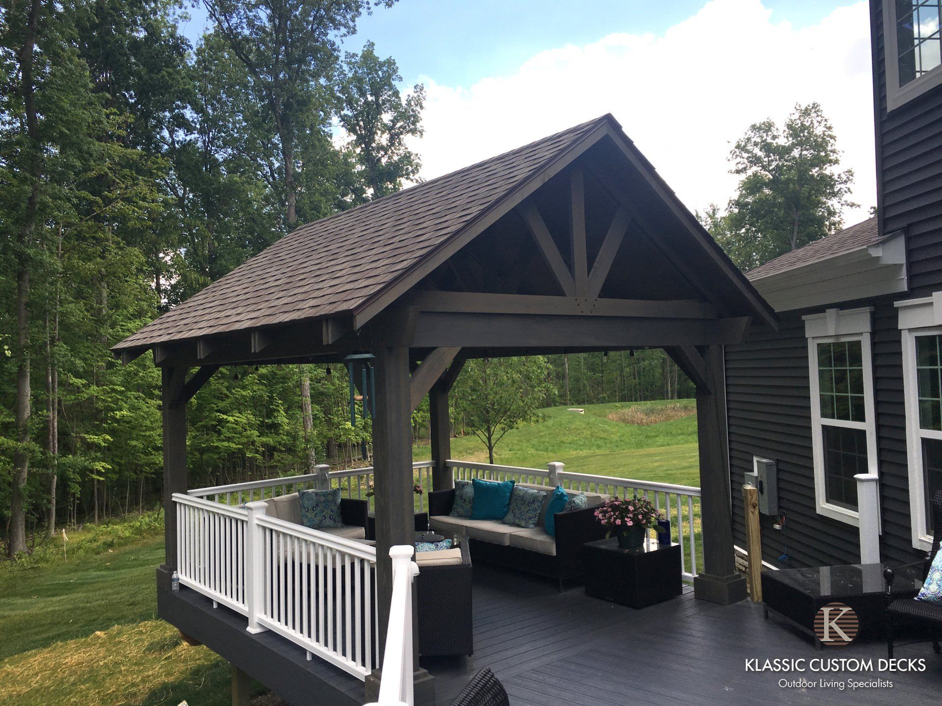 Covered backyard deck with white railings, dark roof, and outdoor seating beside a house and trees.
