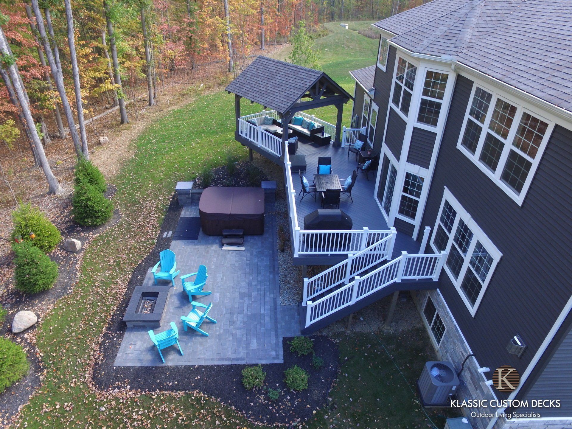 Aerial view of a black deck with blue chairs, grill, gazebo, and stairs beside a house and autumn woods