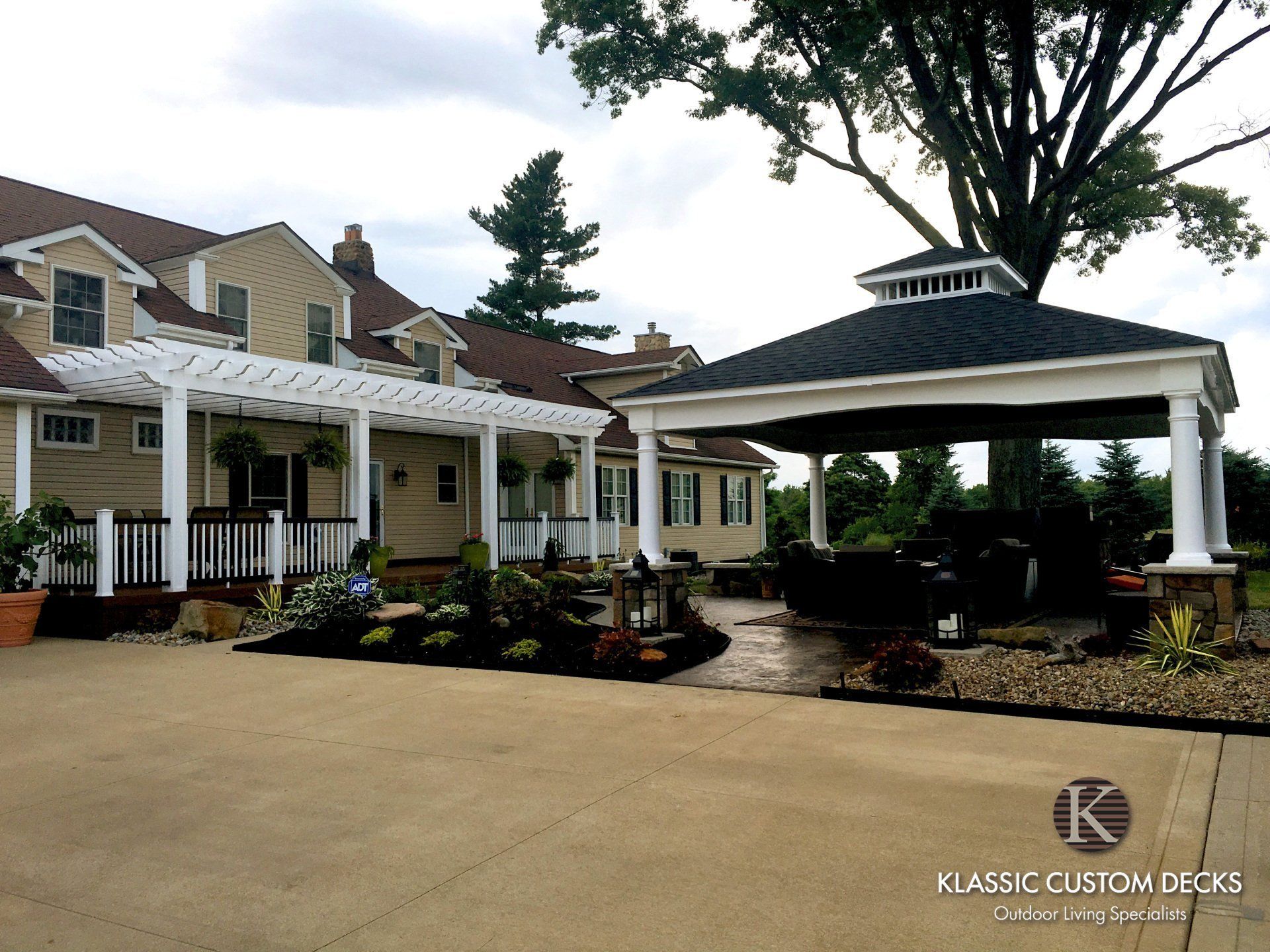 Luxury mansion entrance with white covered driveway, landscaped beds, and large tree under a cloudy sky