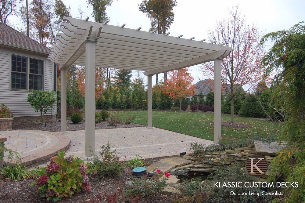 White pergola over a stone patio in a landscaped backyard beside a house