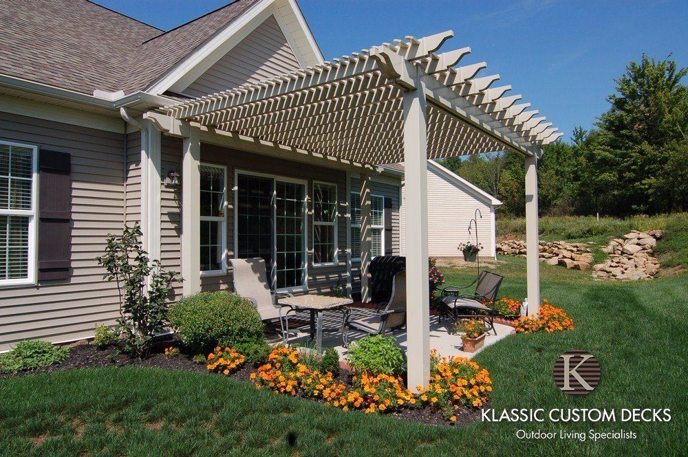 Patio with white pergola beside a house, patio furniture, and colorful flower beds