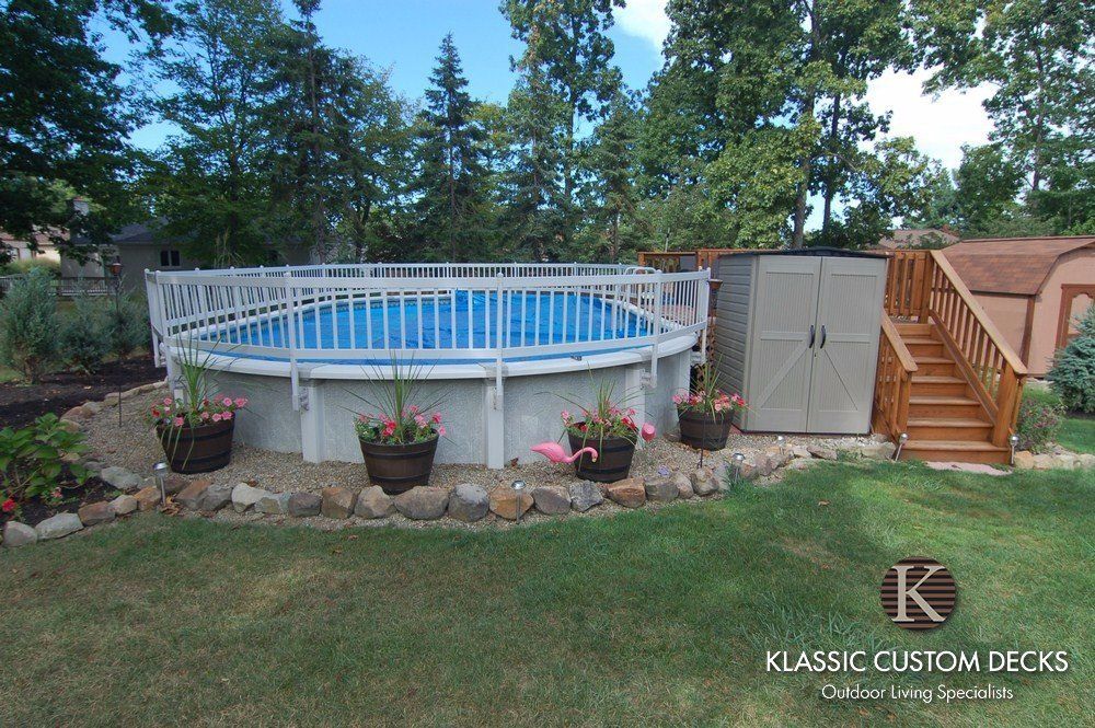 Above-ground pool beside wooden deck and stairs in a backyard with potted plants and trees