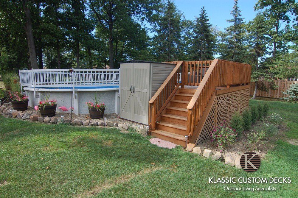 Wood deck with stairs and railing beside a house, bordered by potted flowers and trees