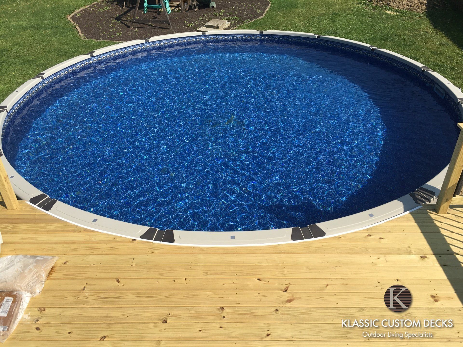 Round blue above-ground pool beside a wooden deck and green lawn in bright sunlight