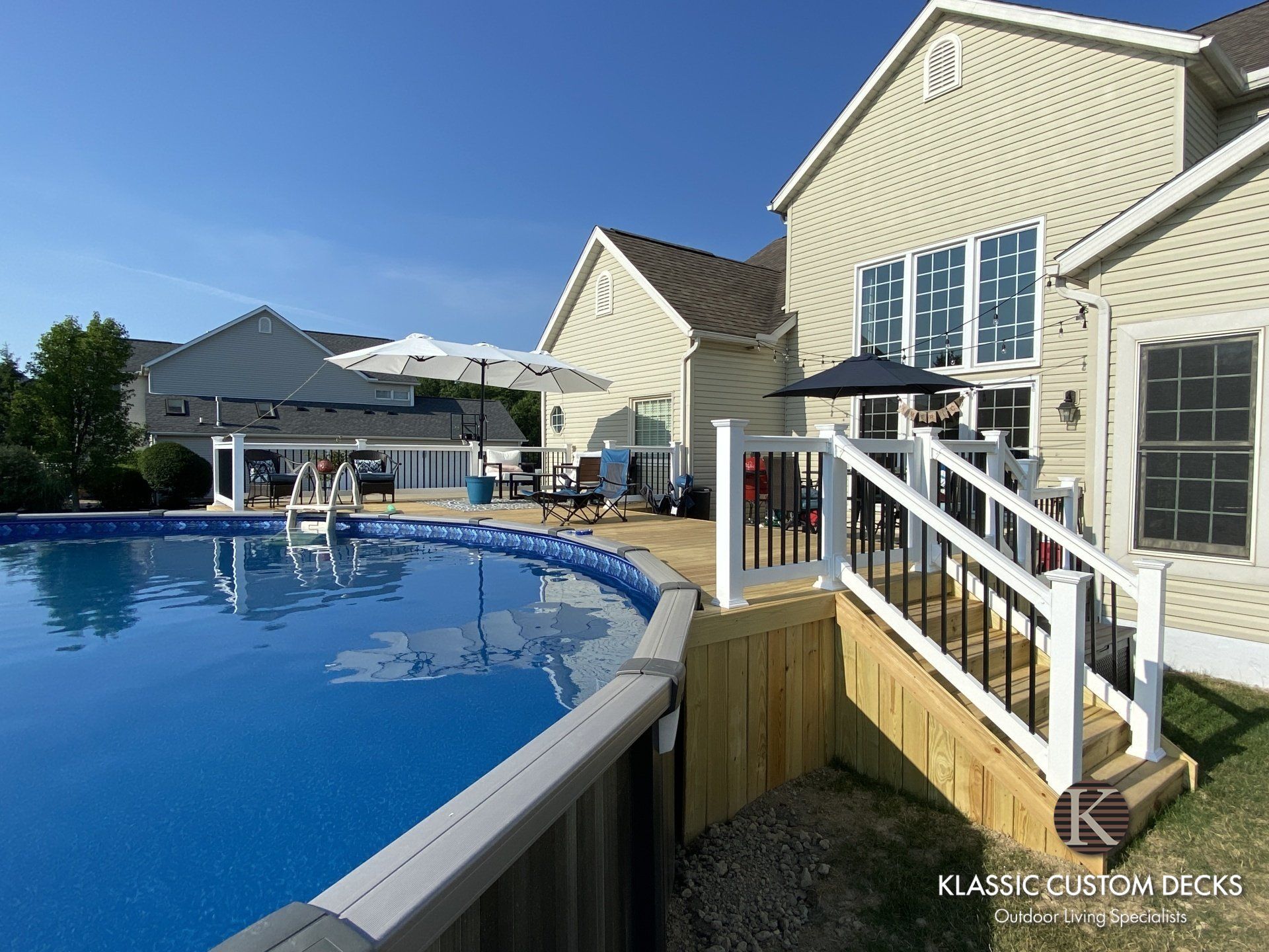 Backyard pool beside beige house and white railing, with patio umbrellas and lounge chairs in sunlight