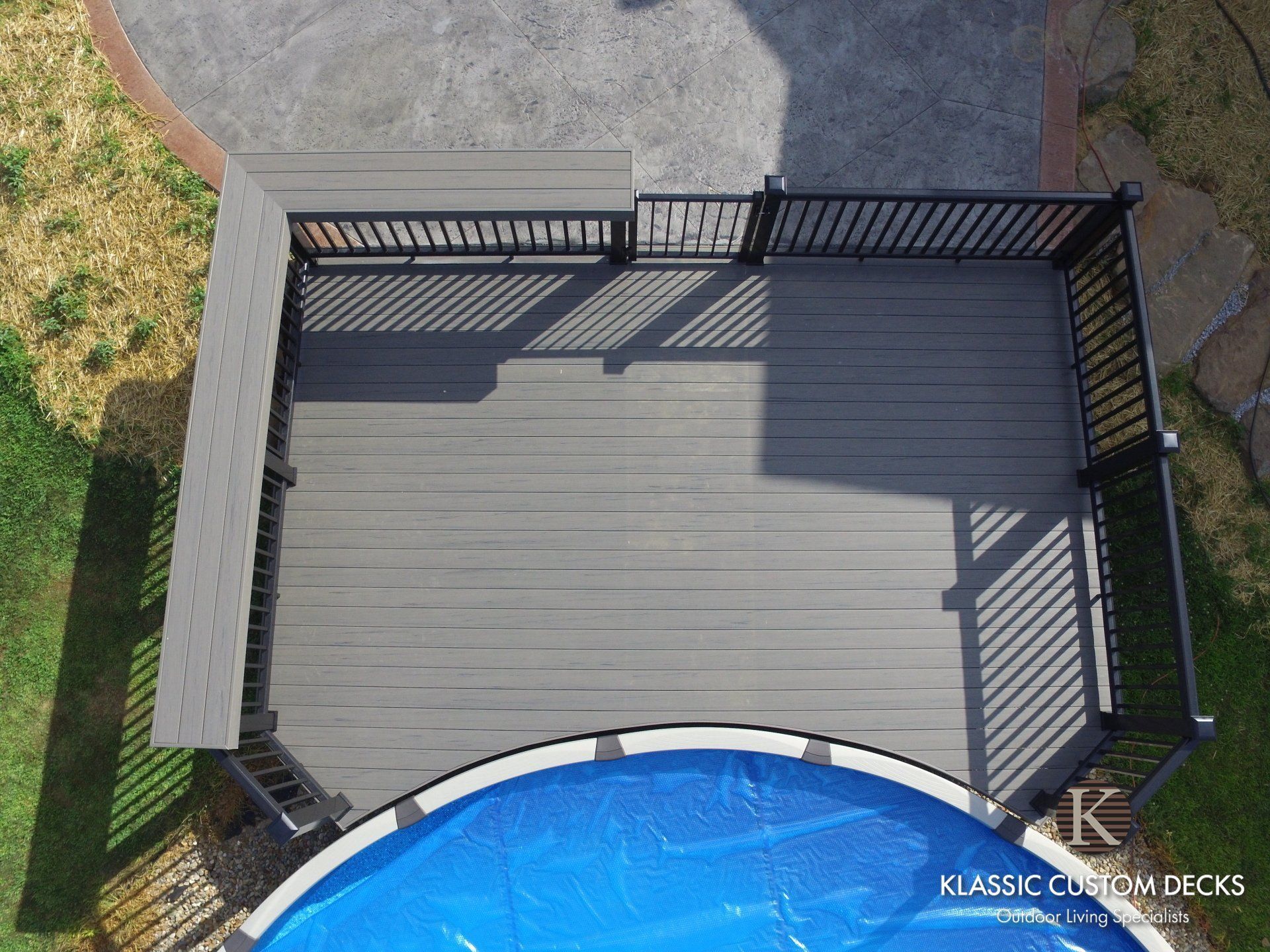 Overhead view of a wooden deck with black railing beside a blue swimming pool and grassy yard