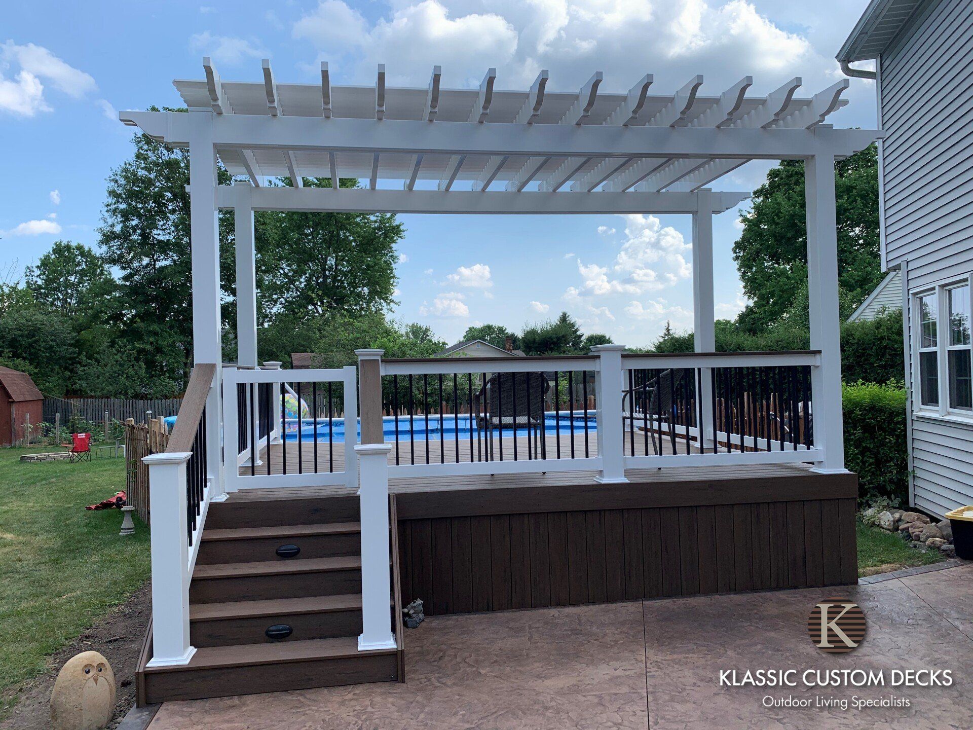 White pergola-covered backyard deck with stairs and railing beside a house and pool