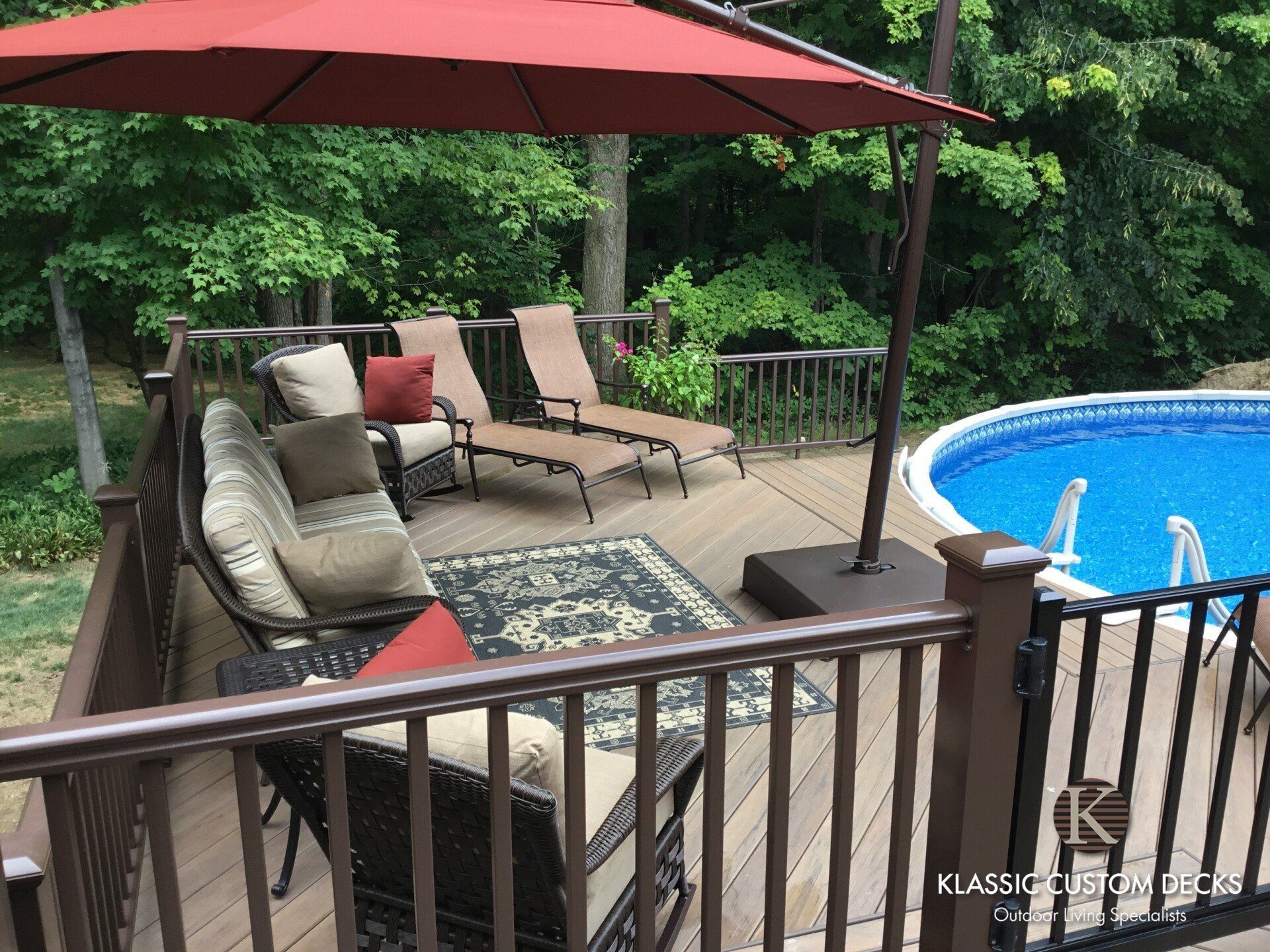 Backyard deck with red umbrella, lounge chairs, and a blue swimming pool surrounded by trees.
