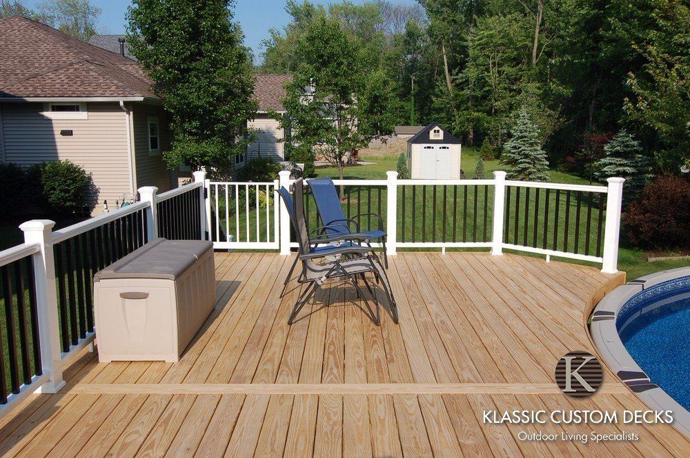 Wooden backyard deck with white railings, tan storage bench, blue chair, and small table in a sunny garden.