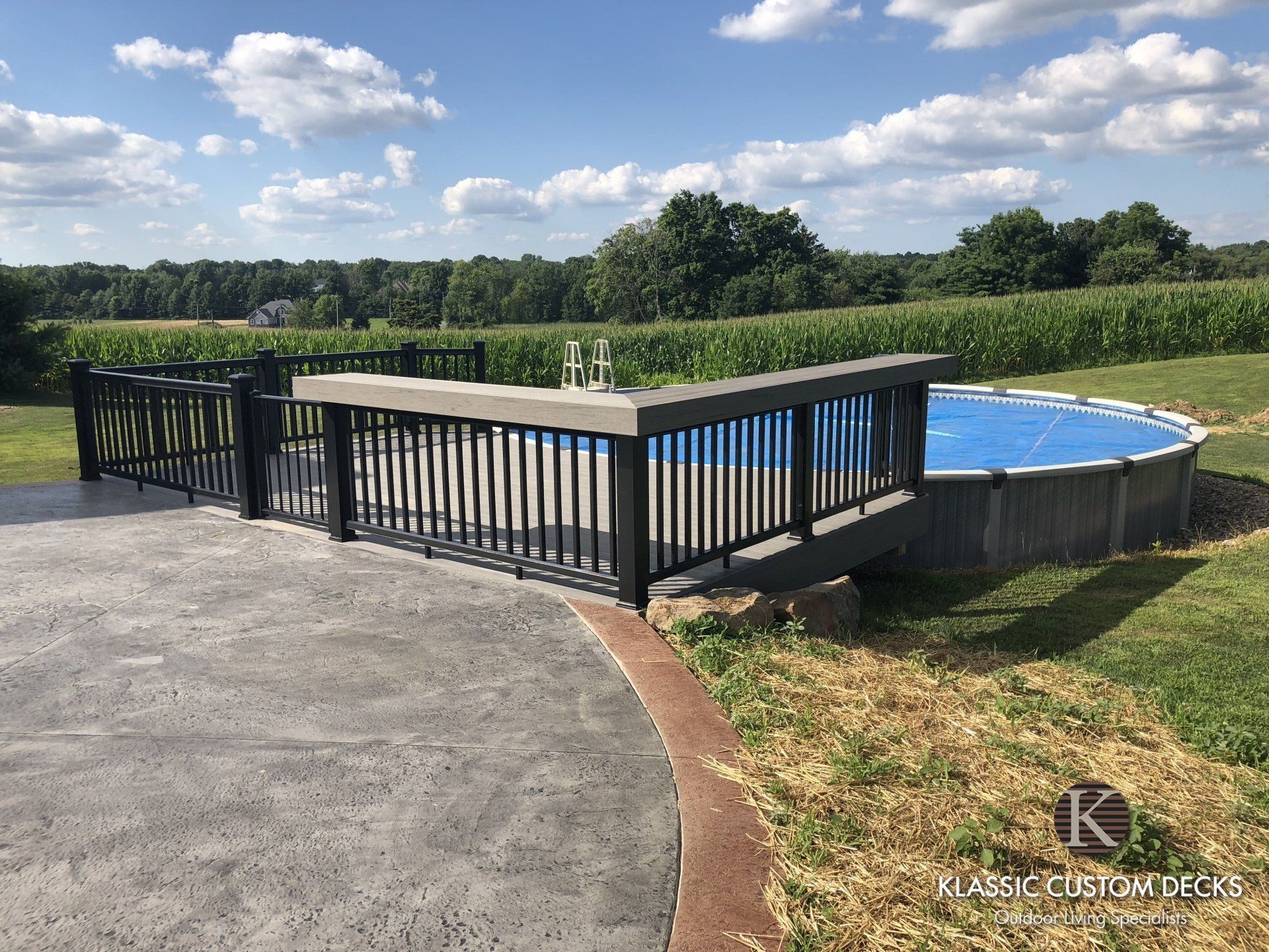 Black fenced overlook with blue pool beside a grassy field under a cloudy sky.