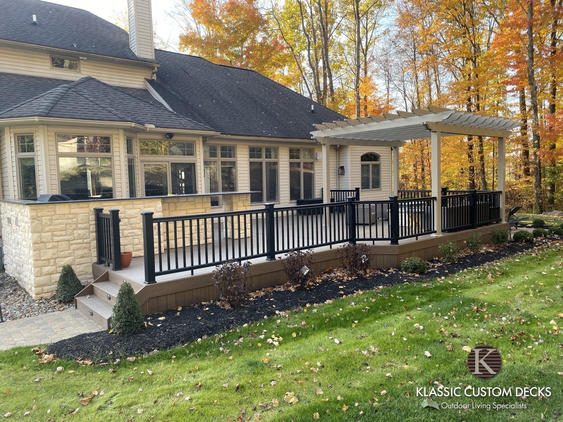 Stone house with dark roof, black porch railing, and autumn trees beside a green lawn