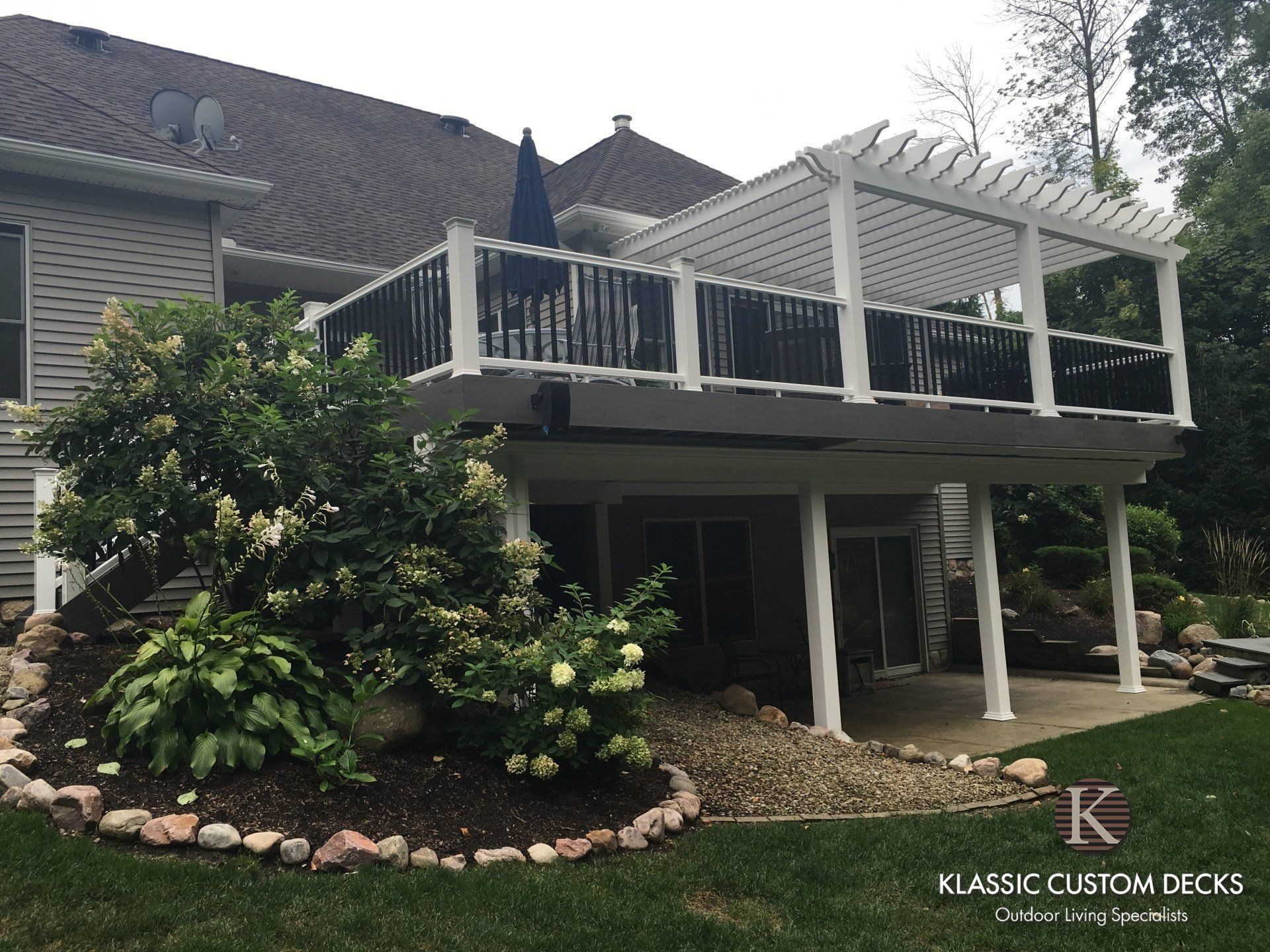 Raised white deck with pergola and stairs beside a house, surrounded by landscaped greenery