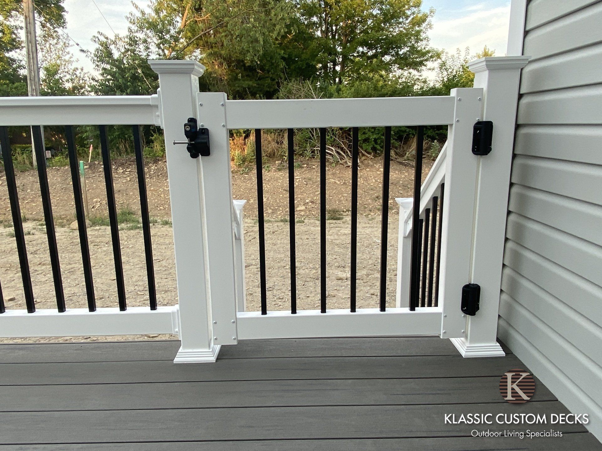 White deck gate with black balusters and hardware, opening to a gravel yard with trees beyond