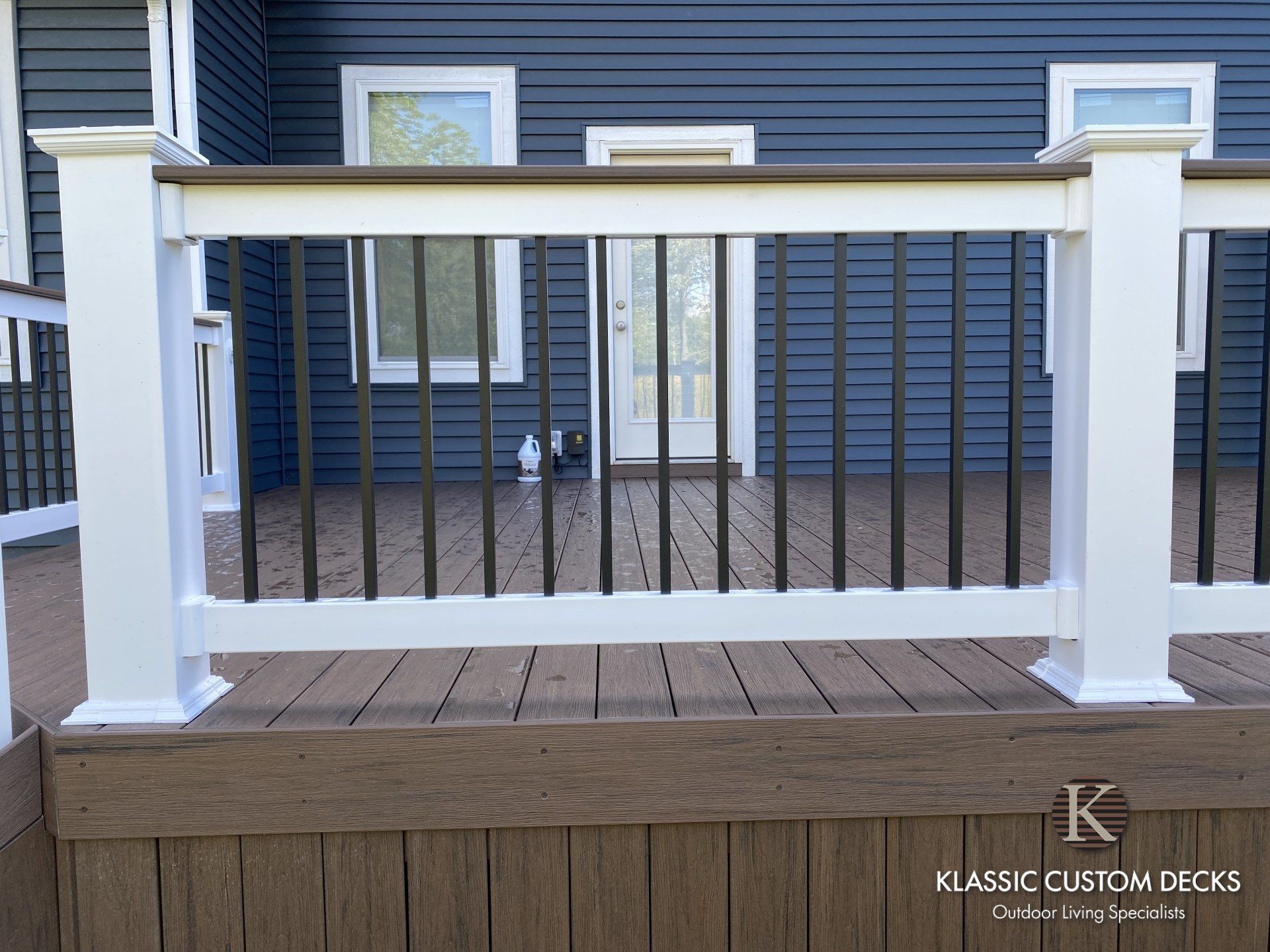 White deck railing with black balusters in front of a blue house, with a small sign on the lower right.