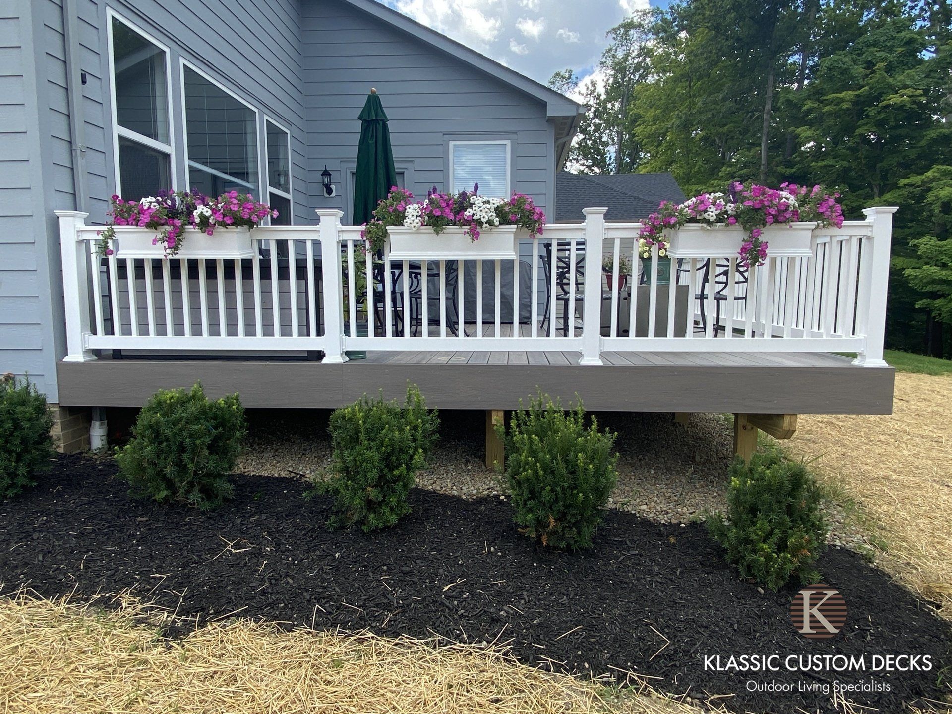 Gray house deck with white railing, pink flower boxes, and shrubs in front of dark mulch.