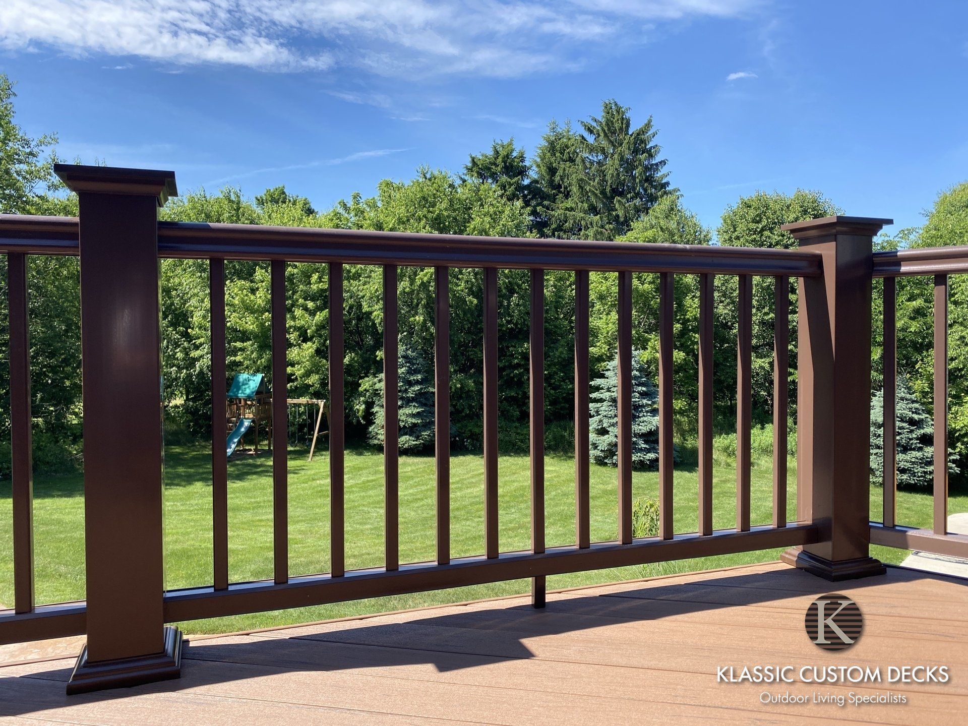 Brown wooden deck railing with vertical balusters overlooking a sunny green yard and trees under a blue sky
