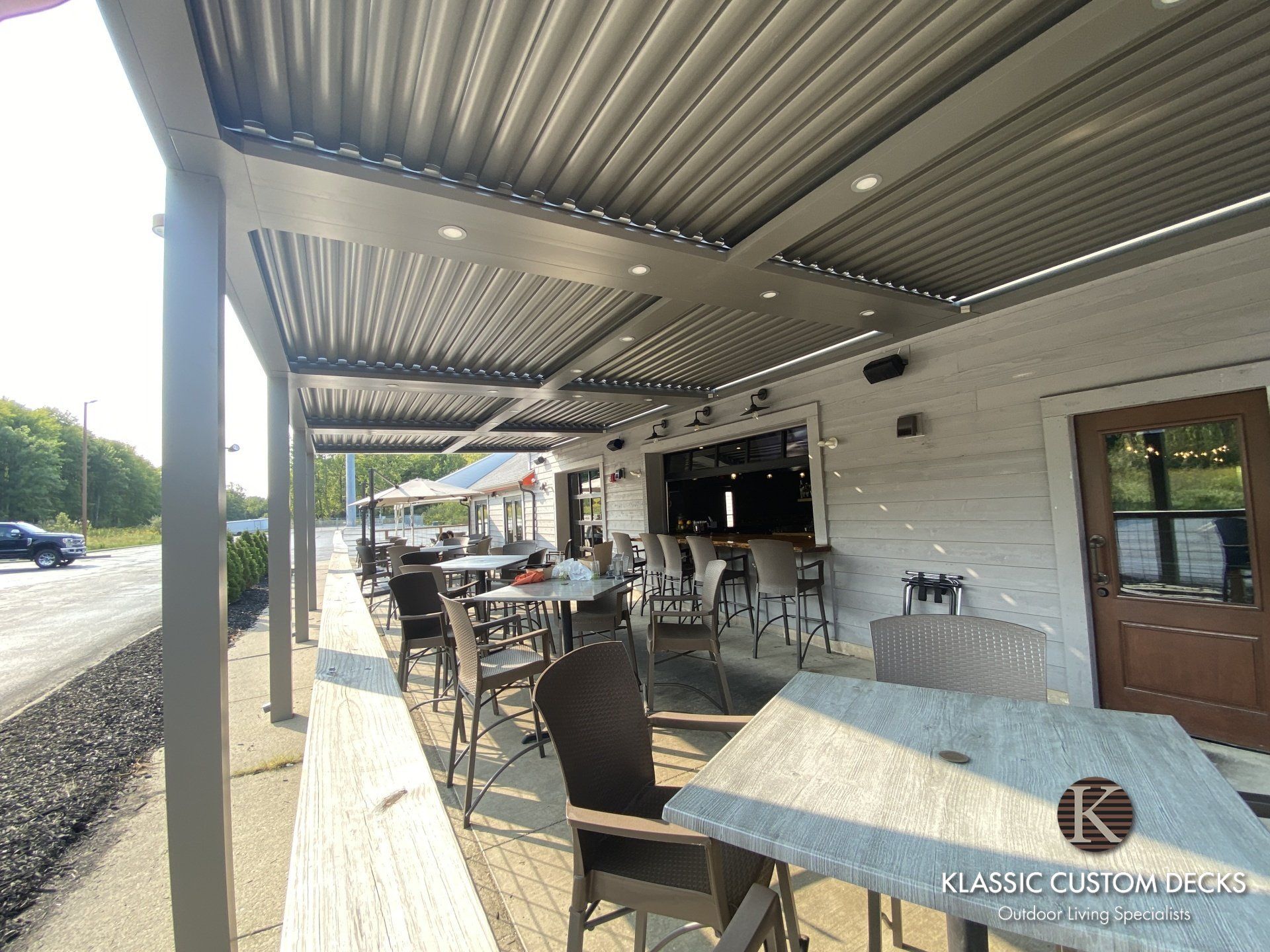 Outdoor café patio with metal chairs, wooden tables, and a covered roof beside a brick wall.