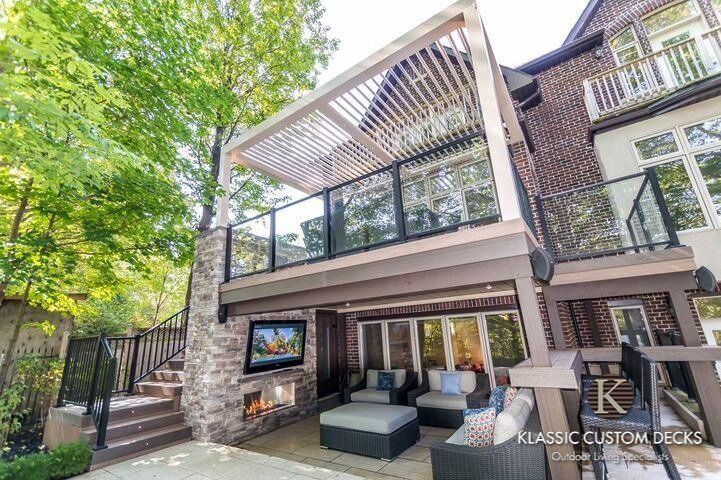 Modern two-story deck with stone fireplace, glass railing, and outdoor seating under a pergola
